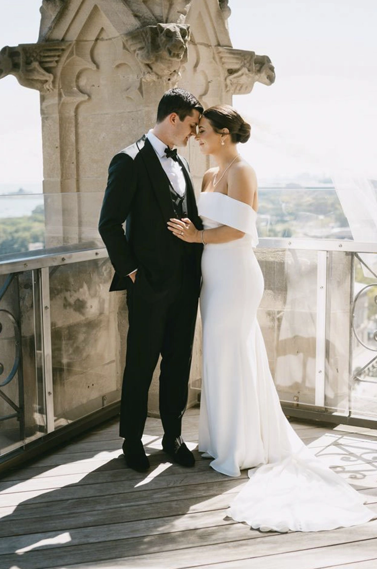 Bride in an off-shoulder crepe gown with a veil and folded over neckline, poses with groom on a sunny rooftop.