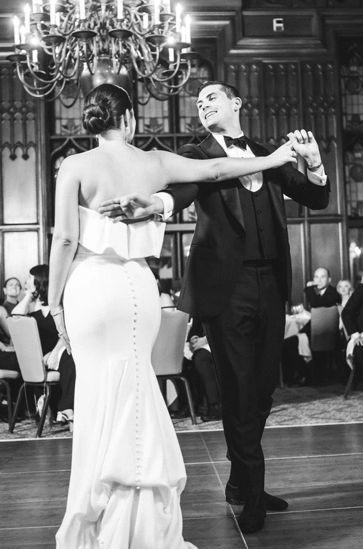 Groom dances with bride in a strapless fit and flare wedding dress with a buttoned back and foldover neckline in a ballroom.