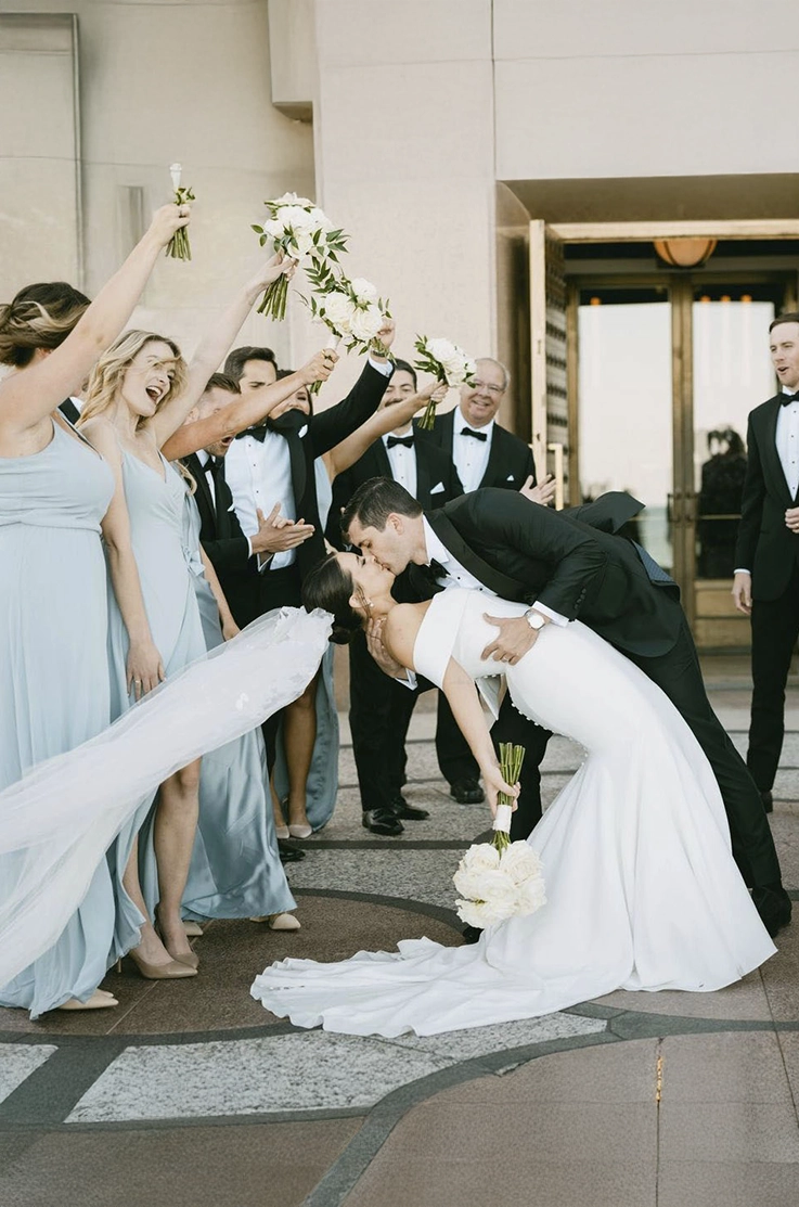Groom dips his bride in an off-shoulder crepe gown with buttons, fit and flare skirt and a veil, as the bridal party cheers.