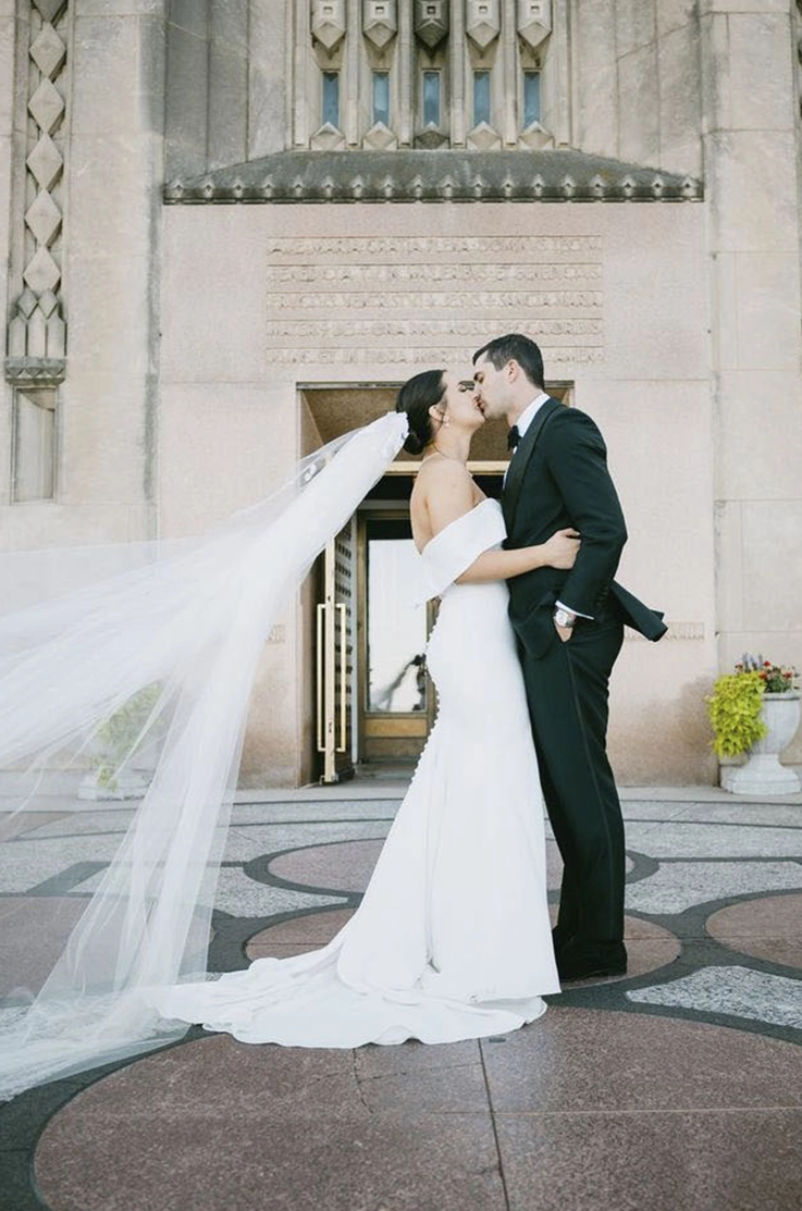 Bride in a crepe fit and flare gown with a portrait collar, buttoned back and veil, kisses groom in front of the venue.