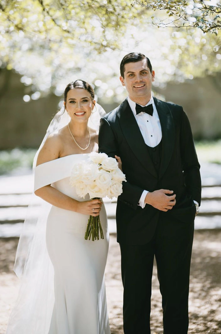 Close-up of bride in a crepe fit and flare gown with a portrait collar and a veil, holding a bouquet beside groom outdoors.