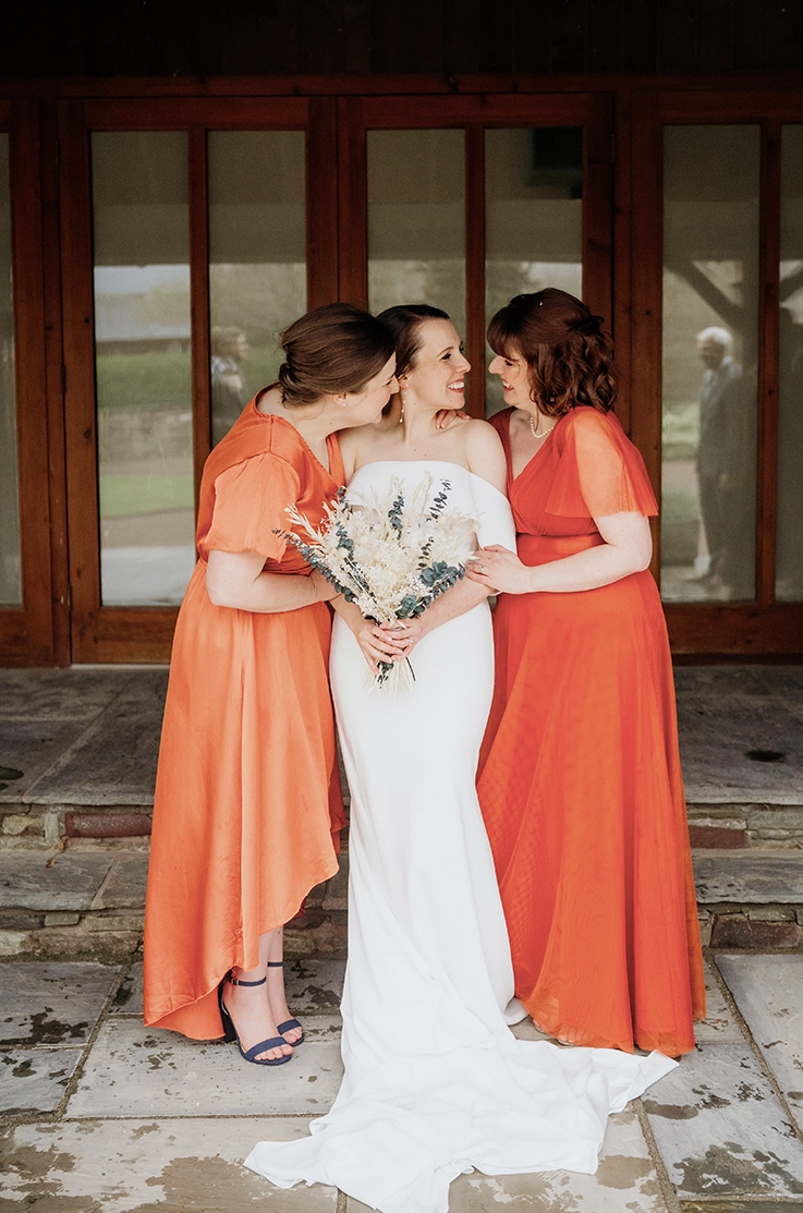 Bride in a strapless off-shoulder crepe wedding dress, holds a bouquet beside bridesmaids in orange dresses outside the venue