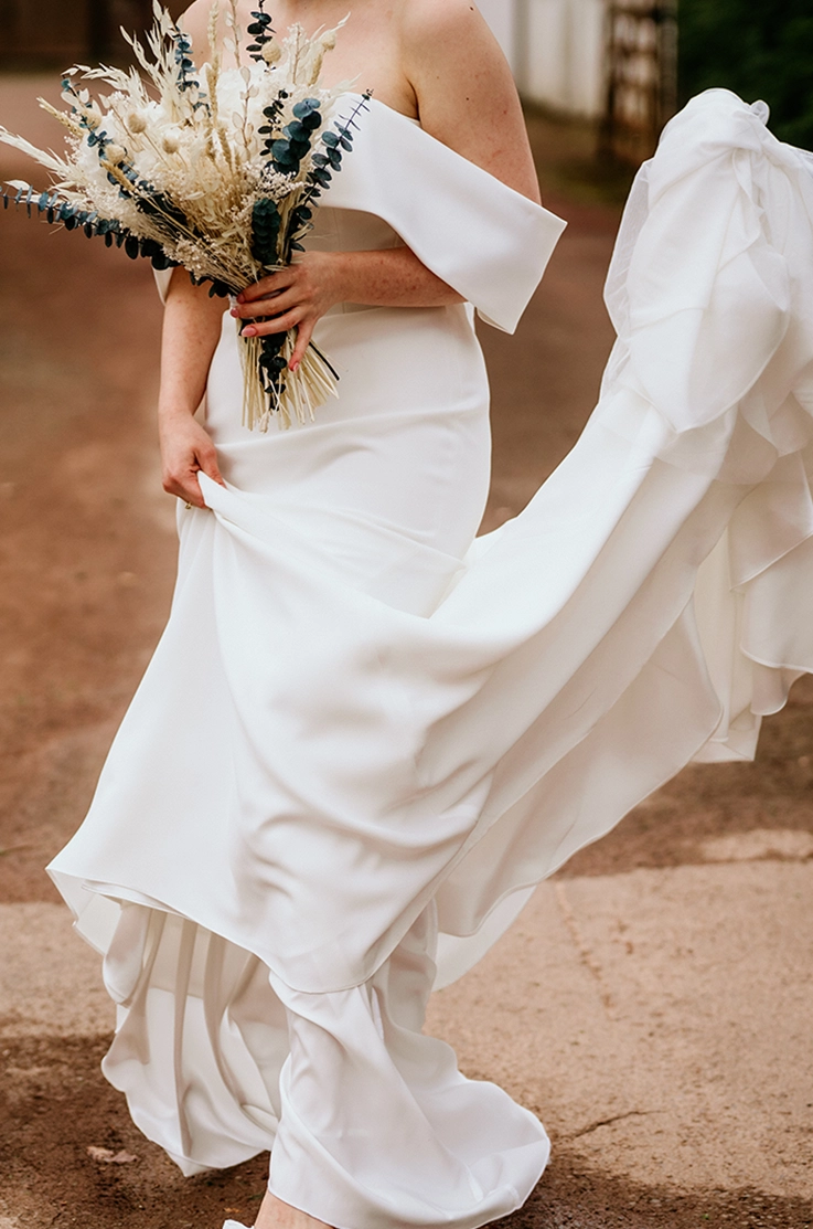 lose-up of bride picking up her train in a crepe fit and flare gown with a portrait collar, holding a dried flower bouquet.
