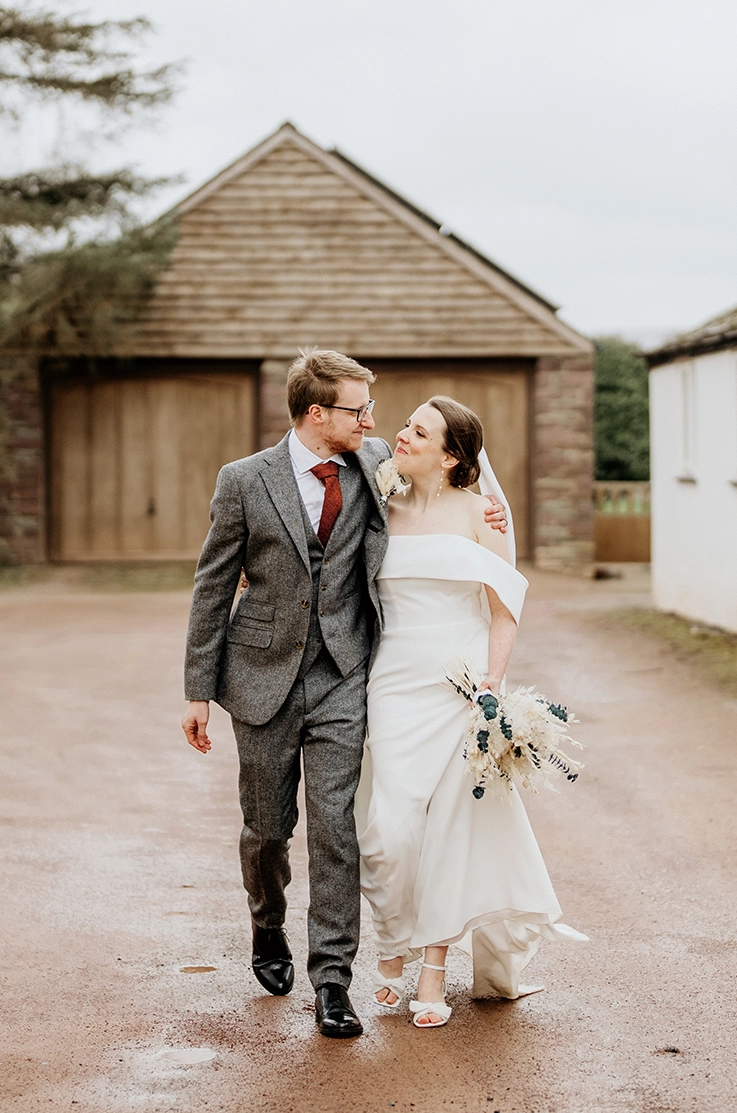 Bride in an off-shoulder crepe fitted gown with a veil ,is holding flowers by a rustic barn outdoors and smiles at her groom.