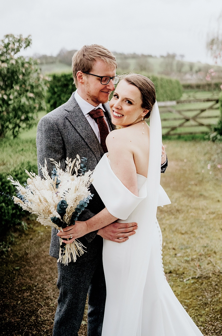 Groom embraces bride in low back crepe fitted gown with a potrait collar and a veil, holding dried flowers in a countryside. 