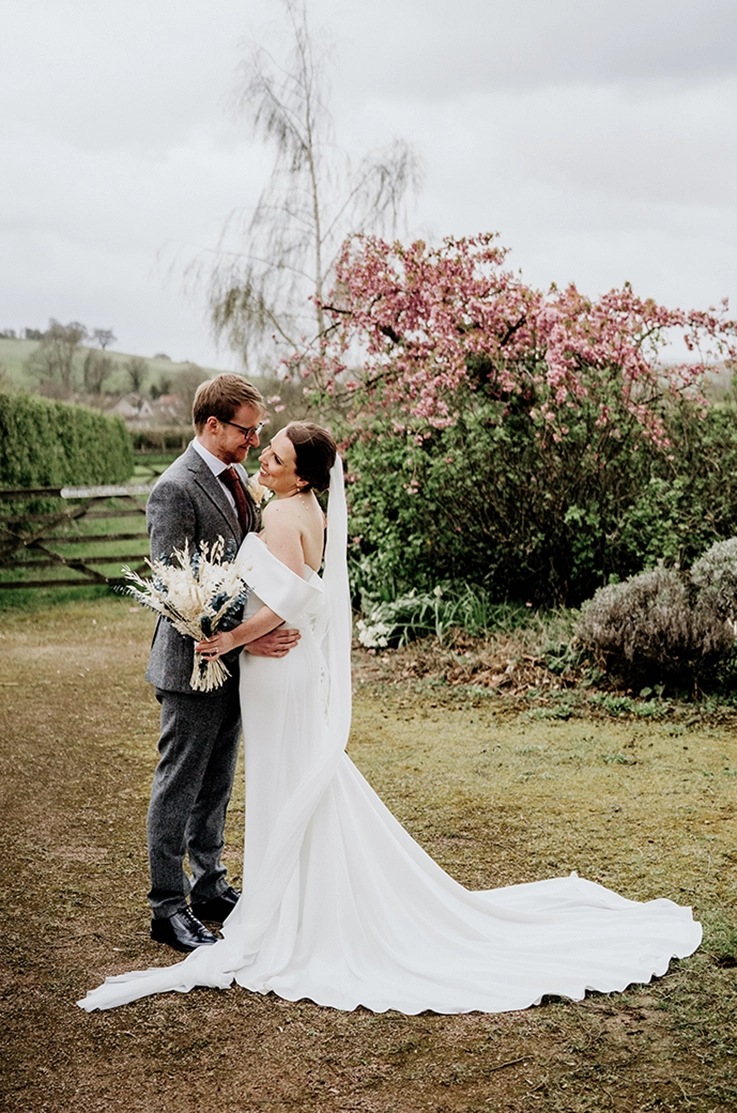 Groom holds bride in an embrace in a off-shoulder crepe fitted gown with a long veil and is holding a dried flower bouquet.