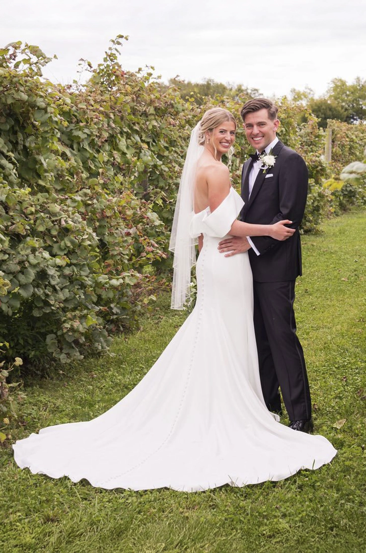 Bride in a fitted off-shoulder gown with buttons and a veil, poses with the groom in a vineyard. 