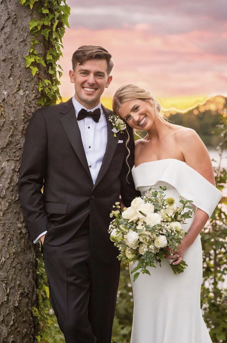 Bride smiles in an off-shoulder crepe fitted wedding dress, holding a bouquet beside groom at sunset by the water.