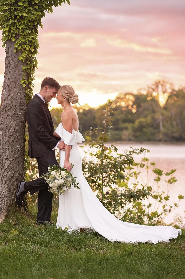 Bride in a crepe fit and flare gown with a portrait collar, holds a bouquet and faces her groom against a tree by a lake.