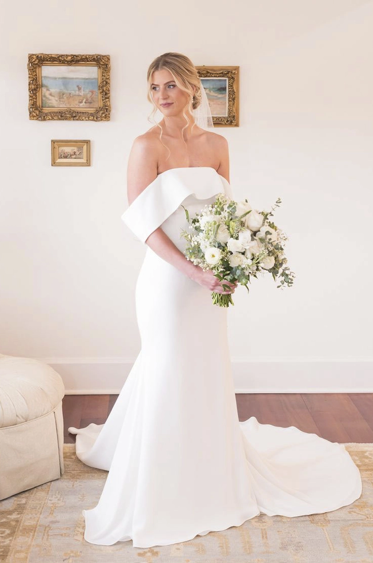 Bride poses in an off-shoulder gown with a crepe fit and flare skirt, standing indoors holding a bouquet by picture frames.