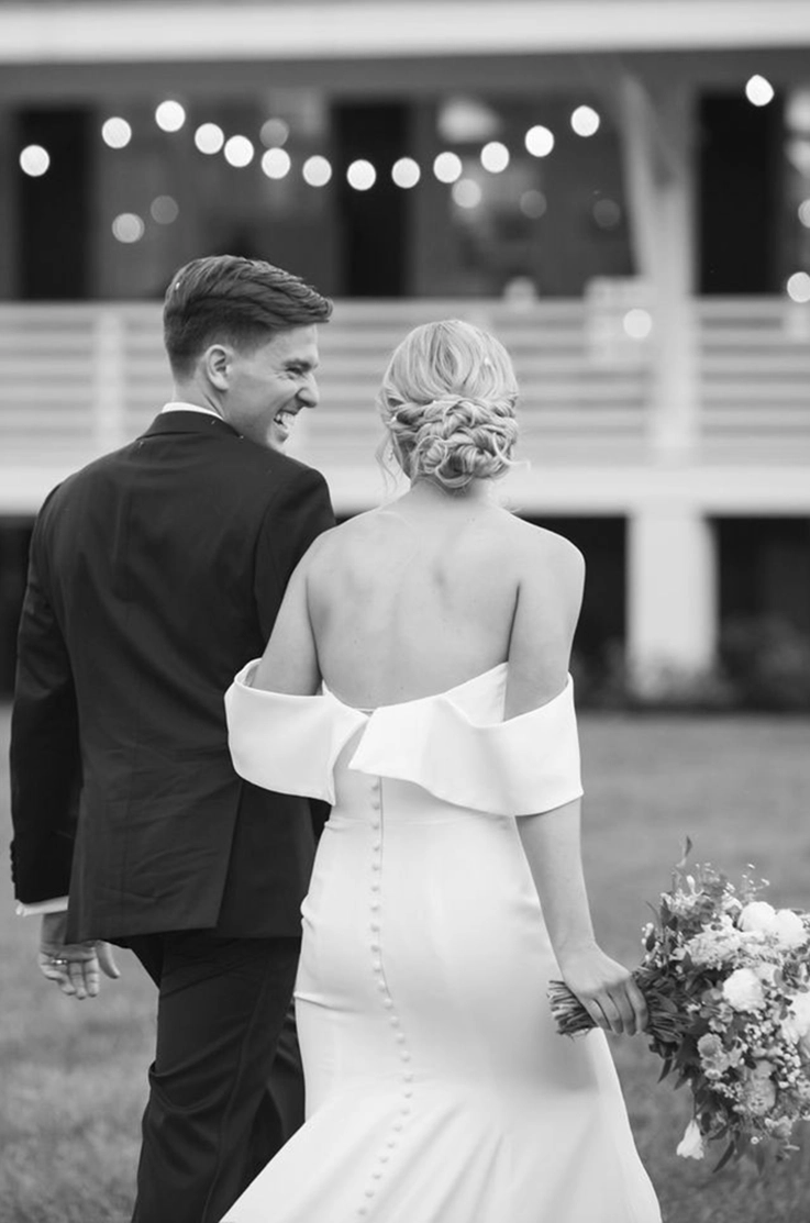 Bride in a crepe fitted gown with a portrait collar and buttons down the back, walks alongside her groom, in black and white. 