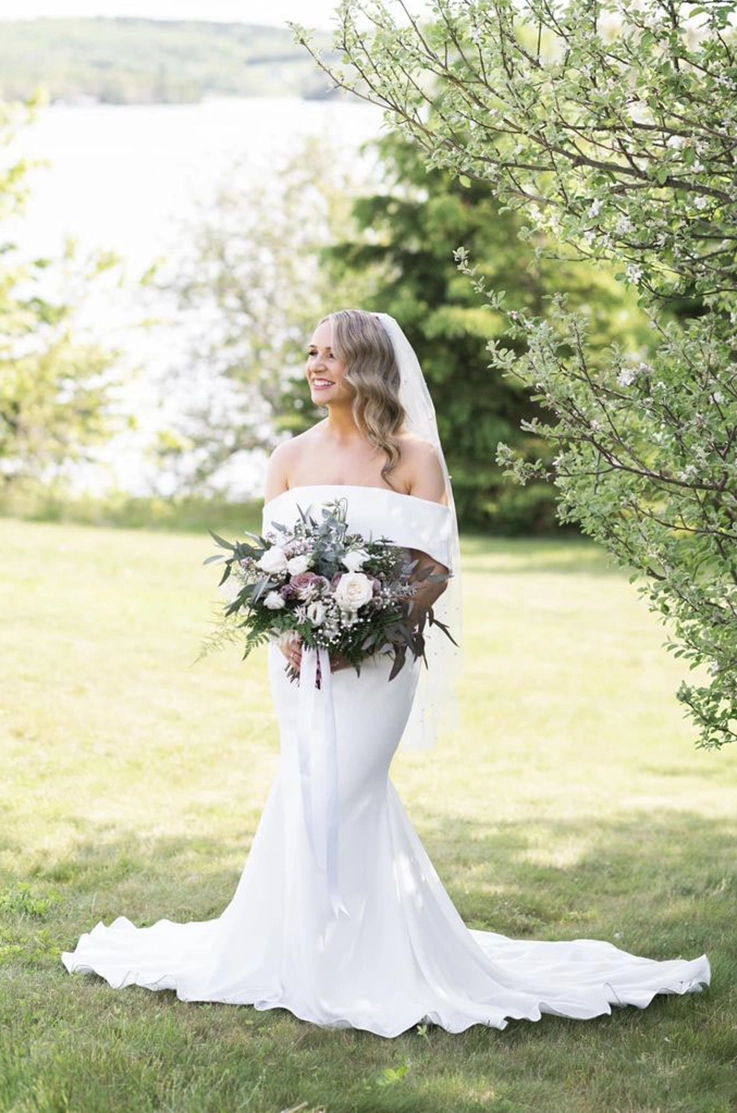 Smiling bride in an off-shoulder fit and flare gown with a veil, is holding a large bouquet in an open sunny field.