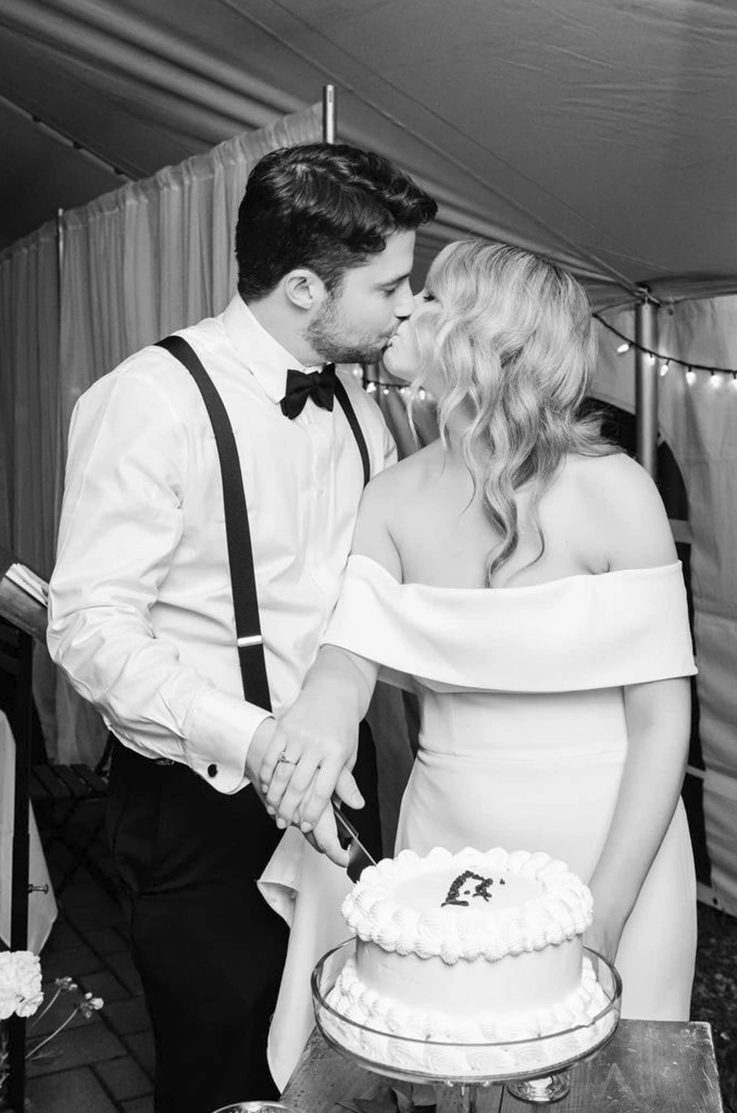 Bride in an off-shoulder fitted gown with a portrait collar, cuts cake with her groom and they kiss, in black and white.