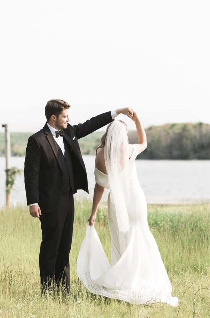 Groom twirls his bride in an off-shoulder crepe wedding dress with a veil and fit and flare skirt, at a lakeside meadow.