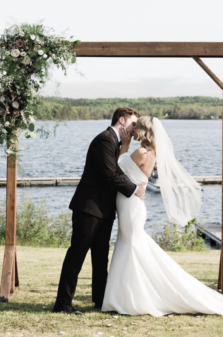 Groom kisses his bride in a strapless crepe fitted gown with a portrait collar and veil, at the altar in waterfront ceremony.