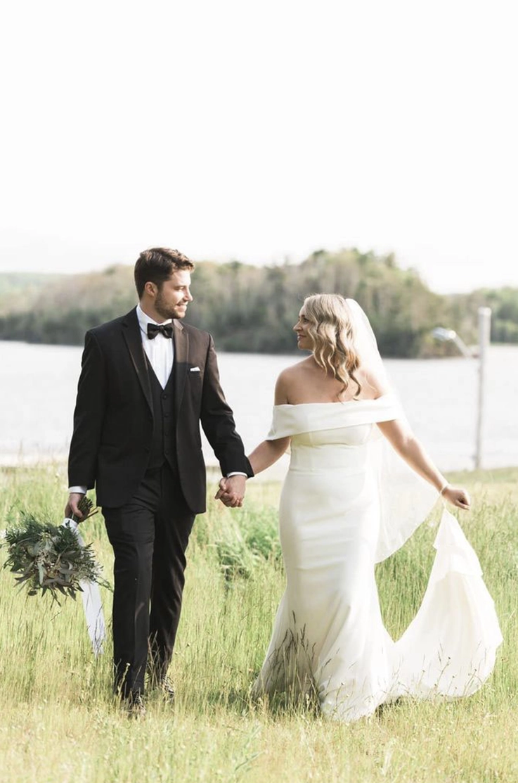 Bride in a portrait collar crepe gown with fit and flare skirt and a veil, walks hand in hand with groom through a meadow.
