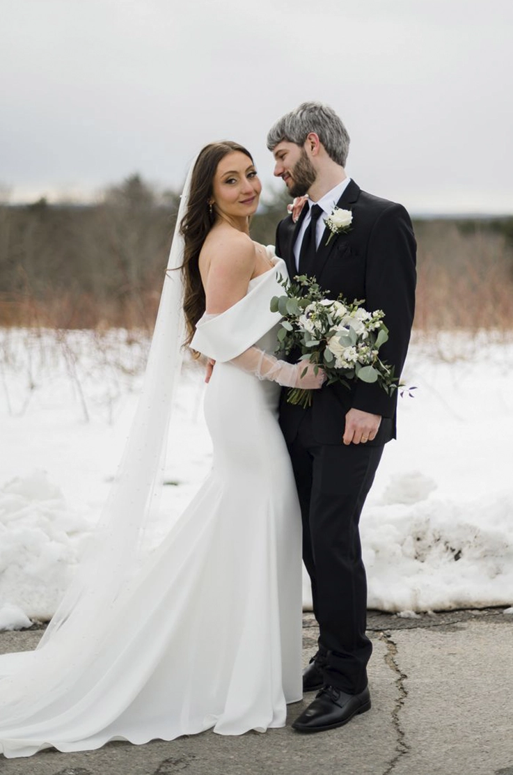 Bride in a portrait collar fit and flare gown with a veil, is holding a bouquet and poses next to her groom on a snowy path.