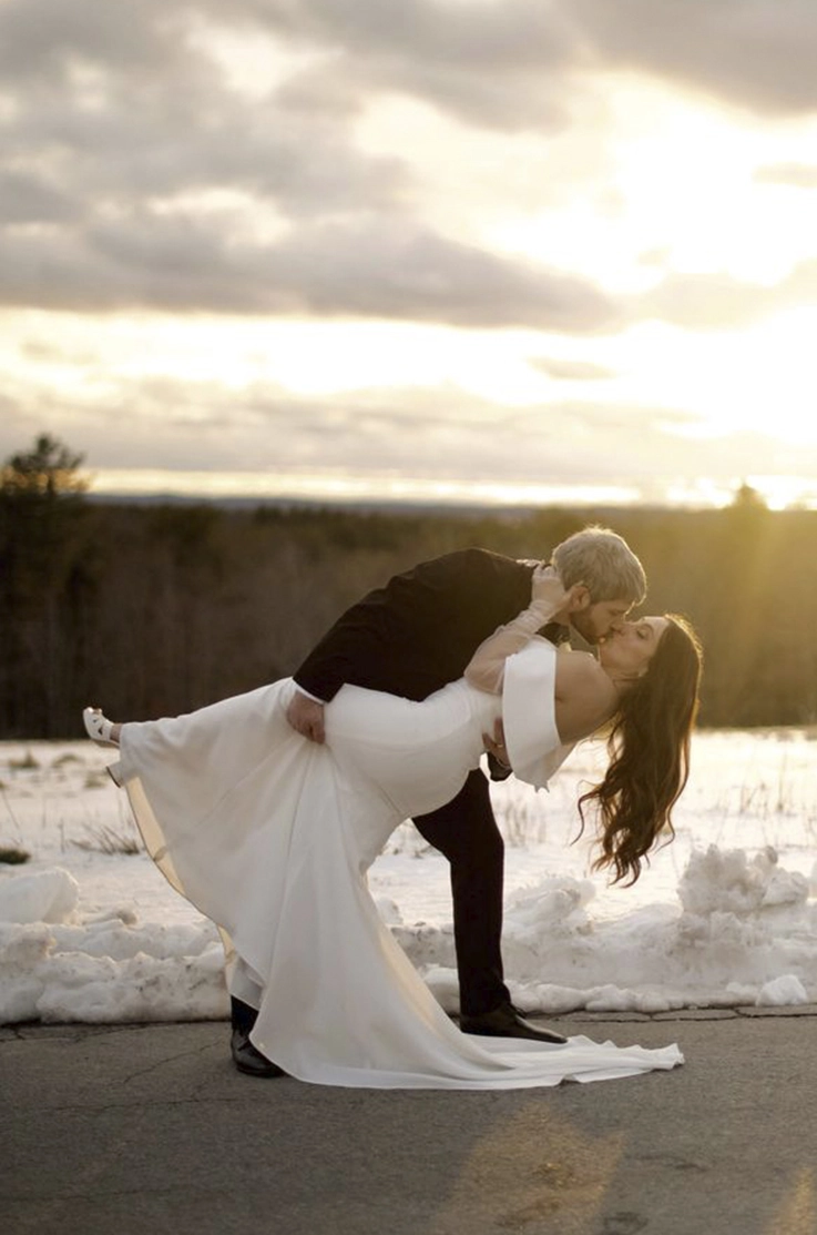 Groom dips his bride in a fitted crepe wedding dress with a portrait collar and they kiss, in winter during a sunset.