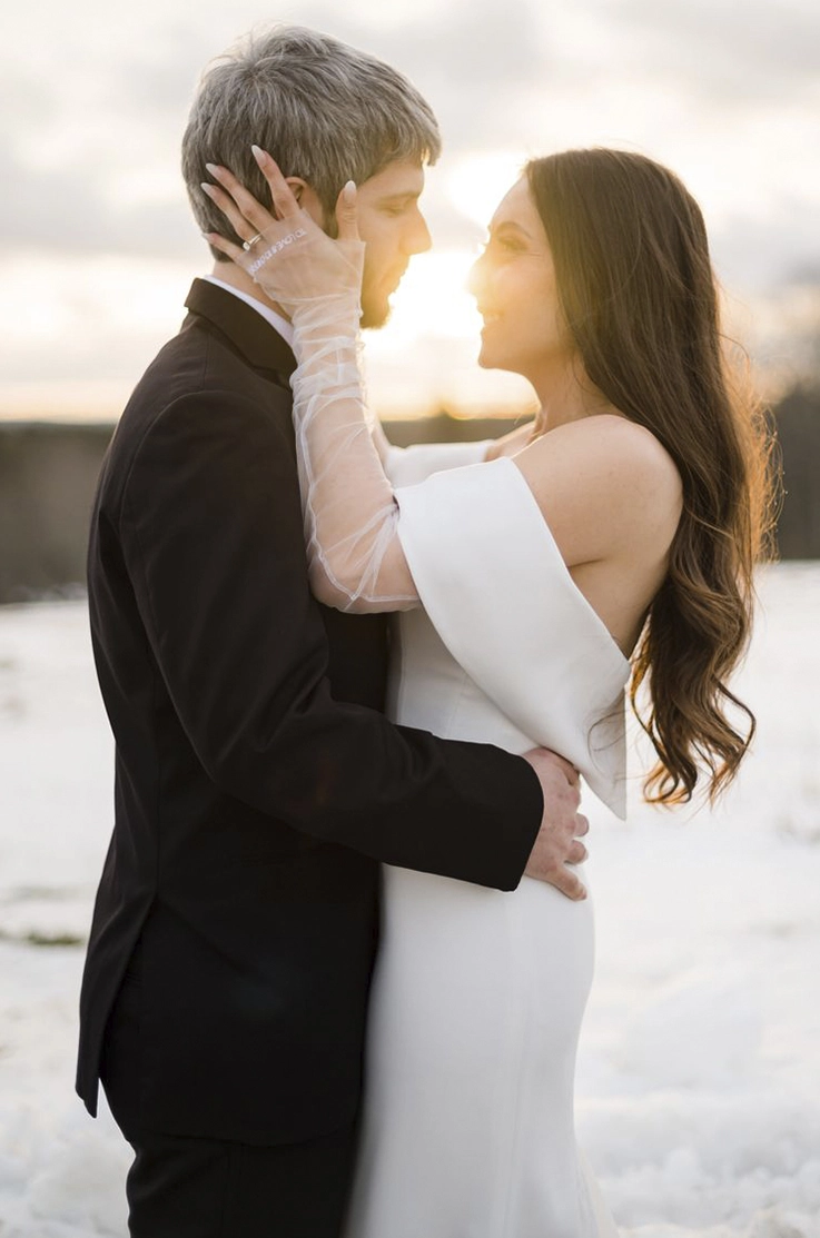 lose-up of bride in an off-shoulder fit and flare gown with sheer sleeves, is facing her groom outdoors in the sunlight