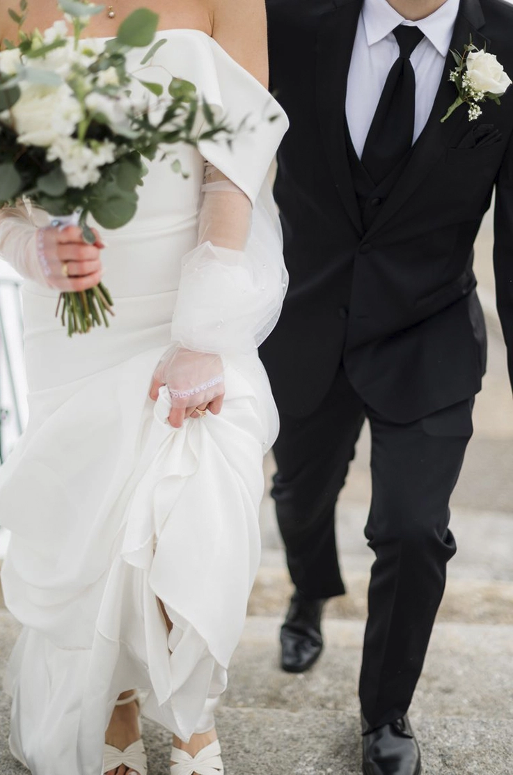 Close-up of bride in an off-shoulder strapless gown with sleeves, picks up her train next to groom while holding flowers.