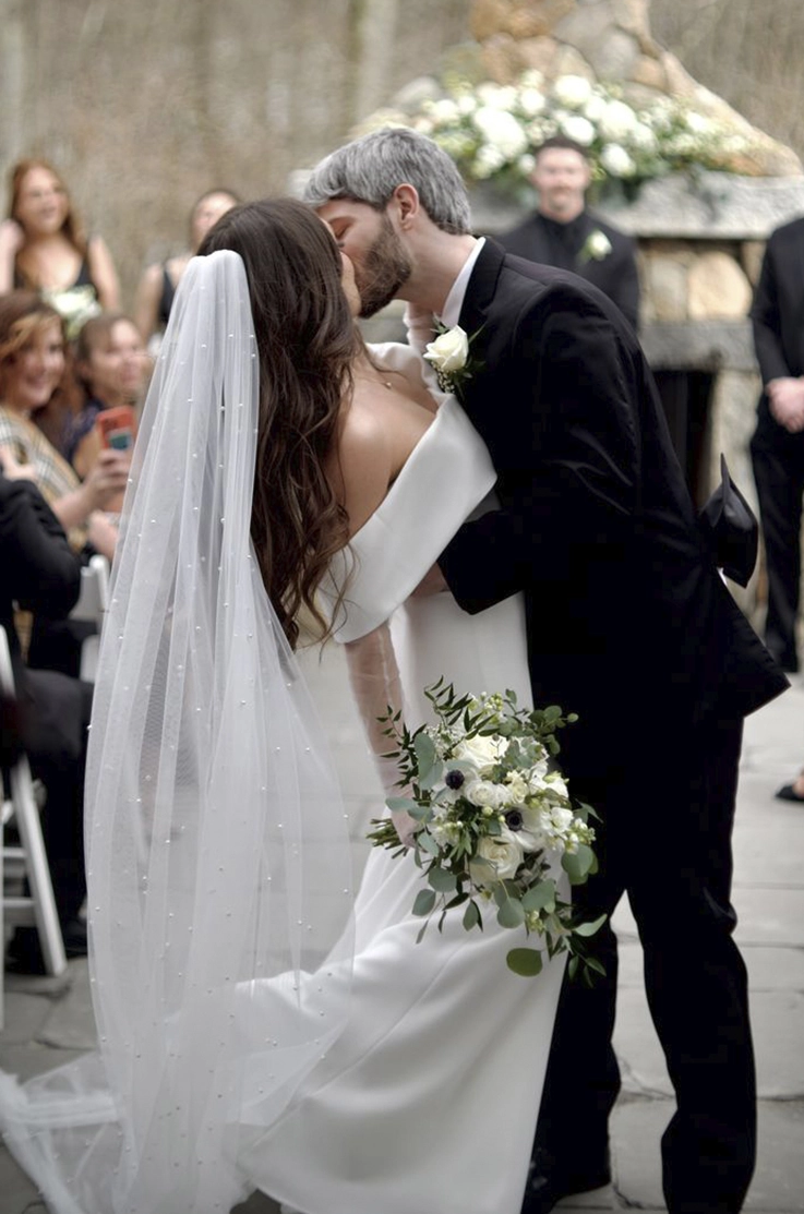 Groom kisses his bride in an off-shoulder crepe gown with sheer sleeves and a pearl-edge veil, in the aisle. 