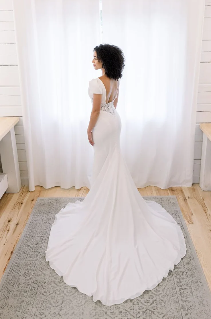 Back of bride posing by a window, in a crepe fit and flare wedding dress with lace detail, a low back and puff sleeves.
