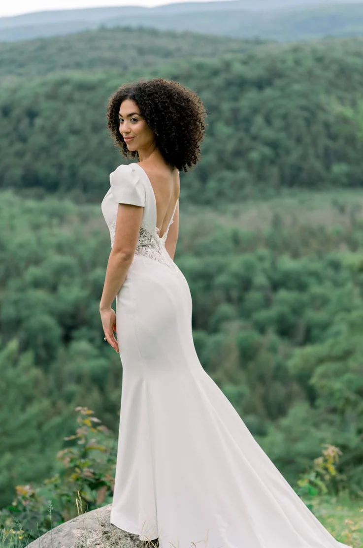 Bride in a cap sleeve crepe and lace gown with an open back and fit and flare skirt, standing on a rock by mountains