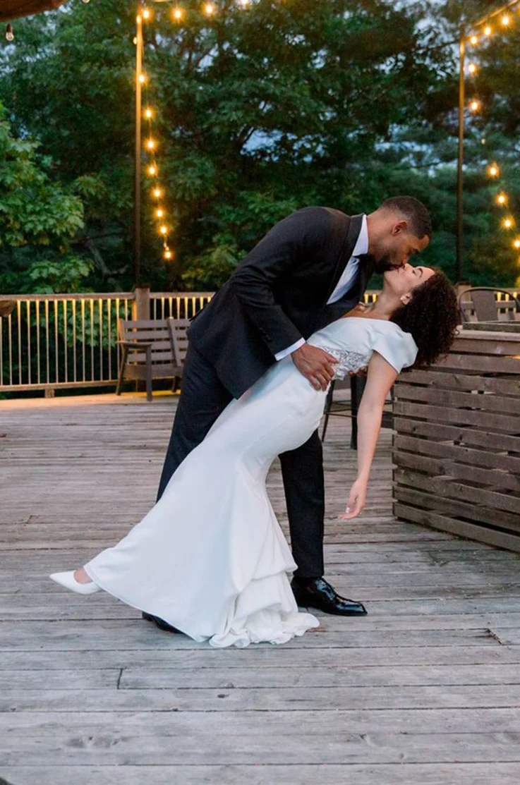 Groom dips his bride in a cap-sleeved crepe gown with lace cutouts and fit and flare skirt, on a deck under string lights.