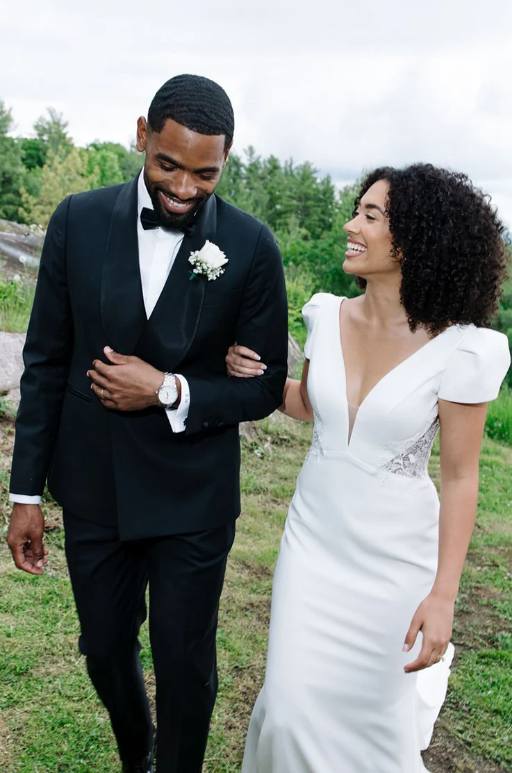 Bride in a V-neck cap sleeve gown with lace on the bodice, walks beside groom and they are linking arms outdoors.