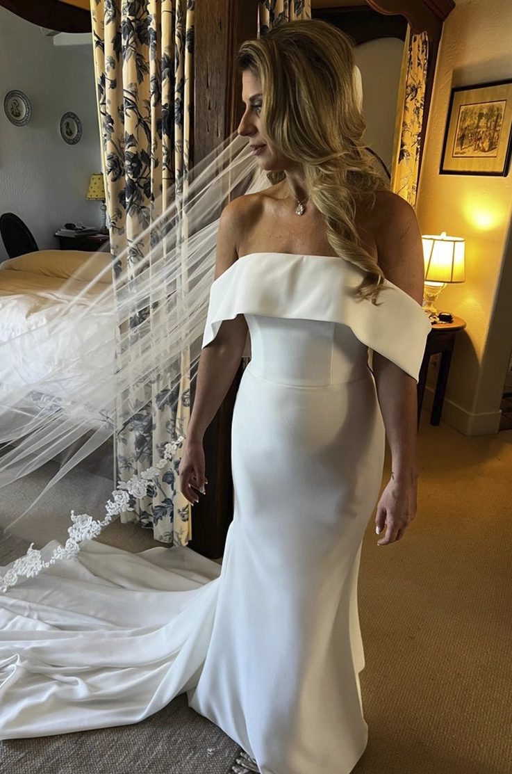  Bride poses in an off-shoulder fitted crepe wedding dress with a lace-trim veil in a bridal suite.