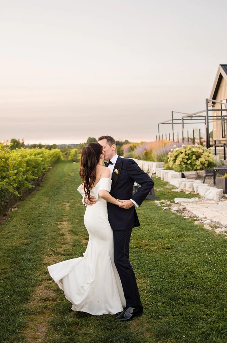 Groom embraces his bride in a fit and flare crepe wedding dress with a portrait collar and they kiss at a vineyard.