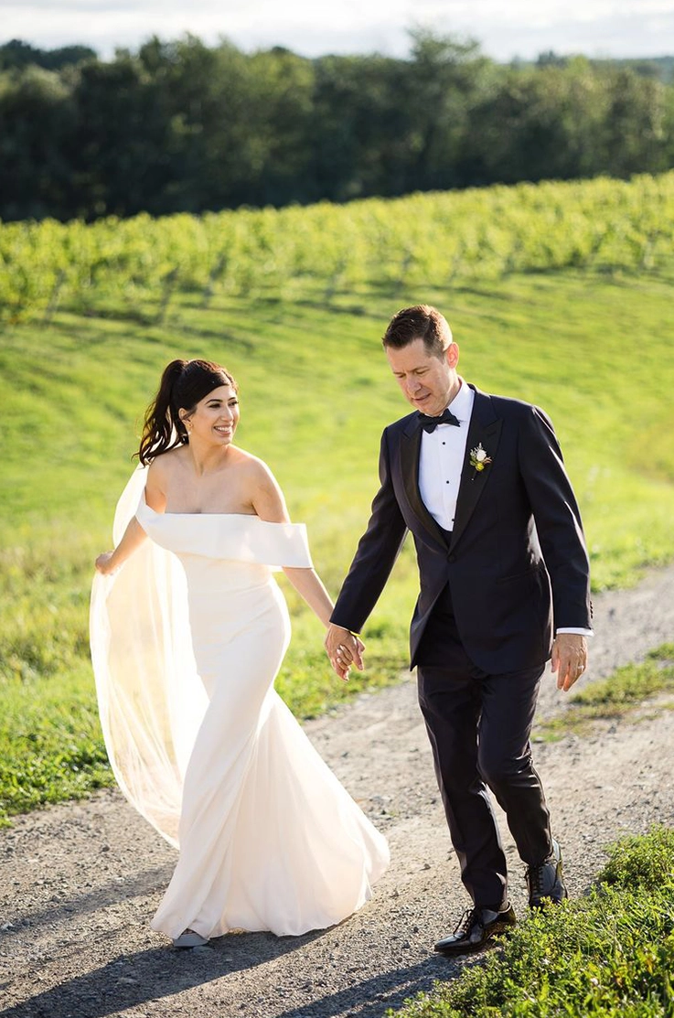 Bride in an off-the-shoulder fitted gown with a flowing veil, walks hand in hand with groom on a sunlight path.