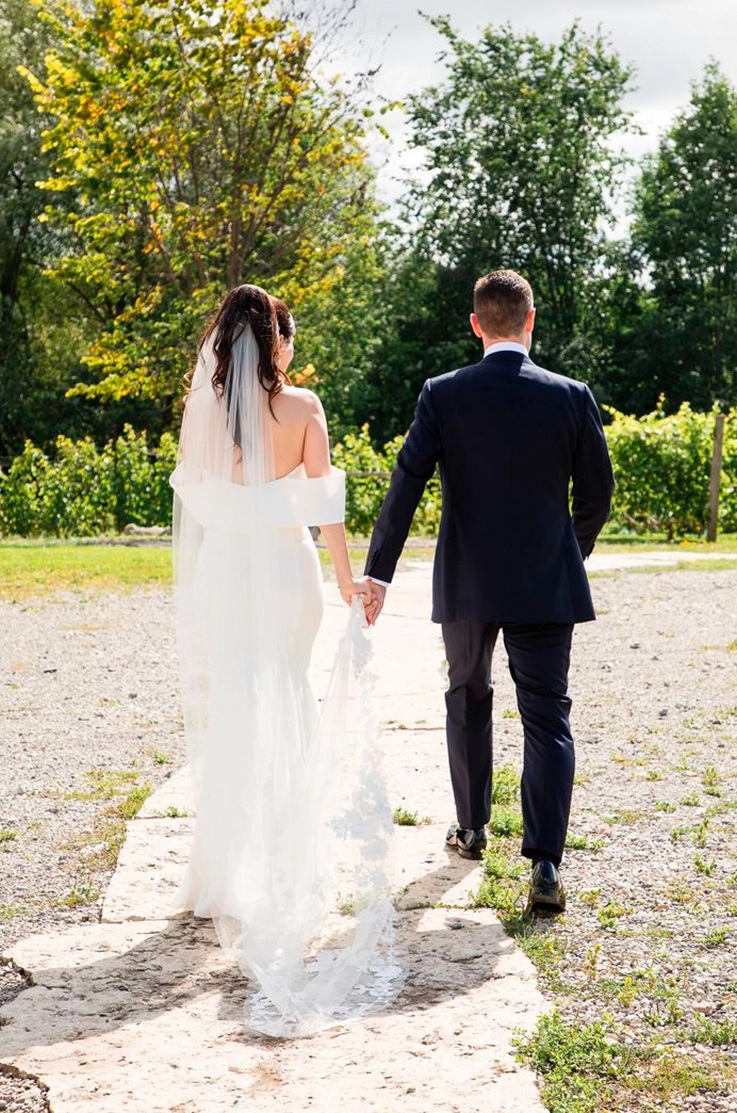 Back of bride in an off-shoulder crepe gown with fit and flare skirt and a lace-trim veil, holds hands with groom outdoors.