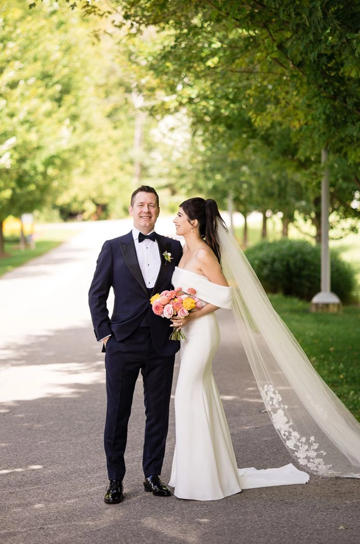 Bride in a crepe off-shoulder fitted gown with a veil, holding a colorful bouquet and poses with groom on a tree-lined path.