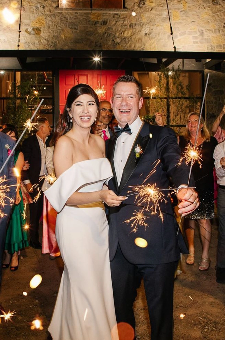 Bride in an off-shoulder fit and flare gown with a portrait collar smiles next to her groom and they are holding sparklers. 