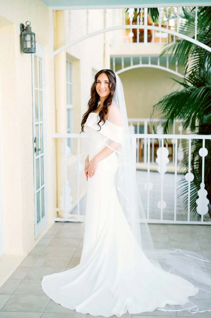 Bride posing in an off-the-shoulder fit and flare gown with a long veil and is standing on an outside porch.