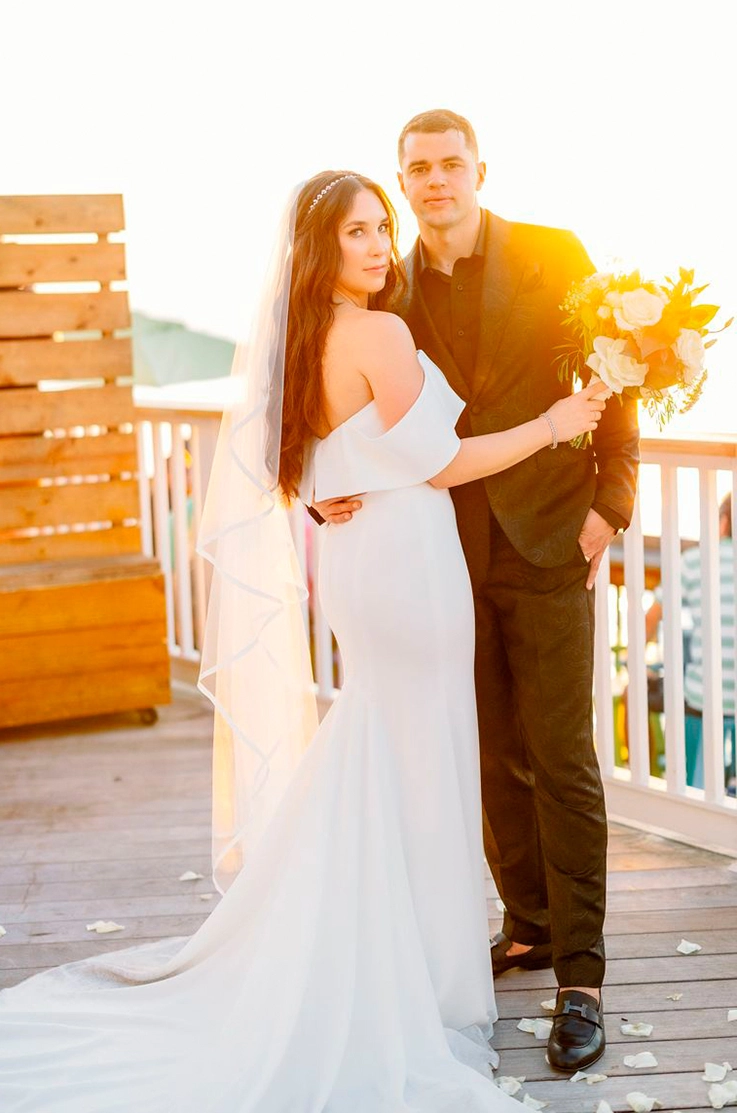 Bride in a crepe fit and flare wedding dress with a long veil and portrait collar, poses with groom on a terrace at sunset.