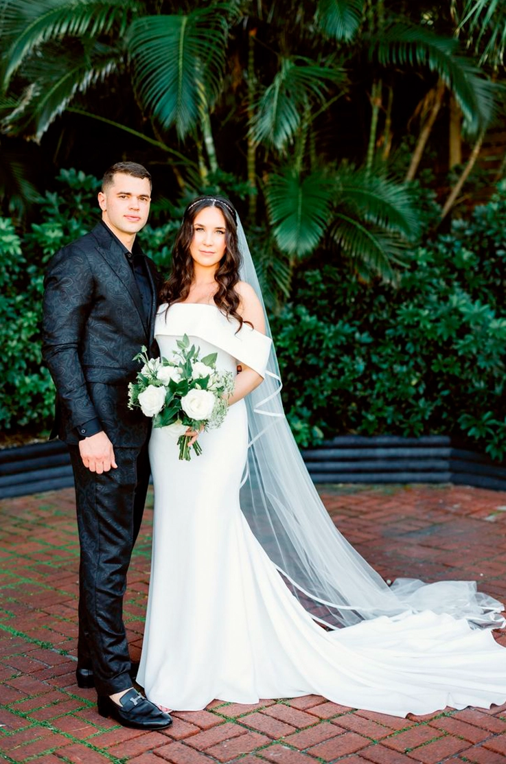 Bride in an off-shoulder crepe gown with fit and flare skirt and a veil, holding a bouquet next to groom under palm trees.