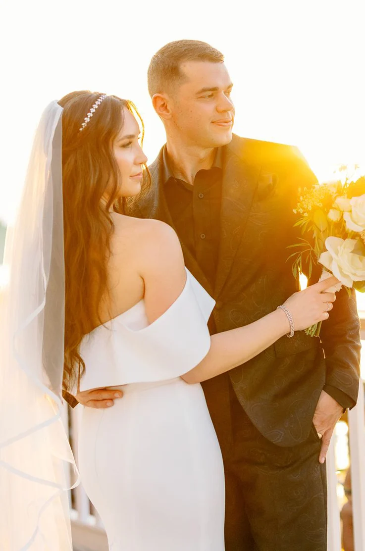 Close-up of groom embracing bride in an off-shoulder fit and flare gown with a veil, holding a bouquet as the sun sets.