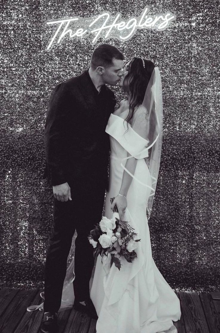Bride in an off-the-shoulder fitted gown with a portrait collar and veil, kisses groom at a backdrop, in black and white.