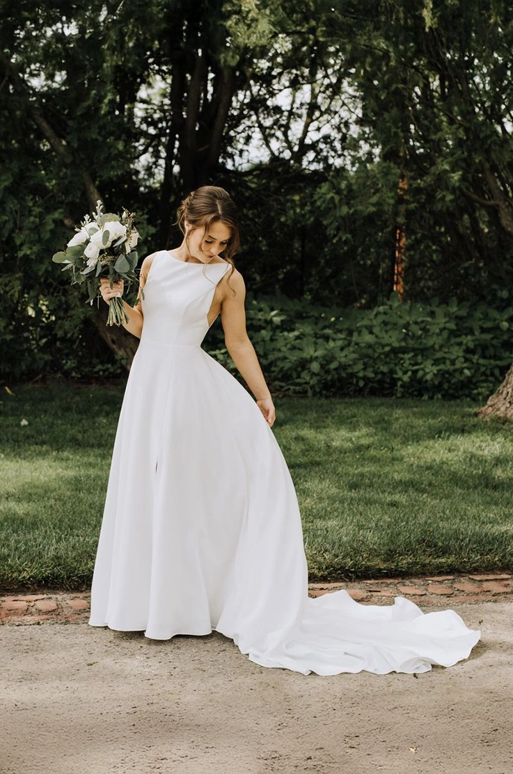 Bride in a sleeveless crepe gown with a bateau neck, is holding a bouquet  and lifts up her train on a garden path.