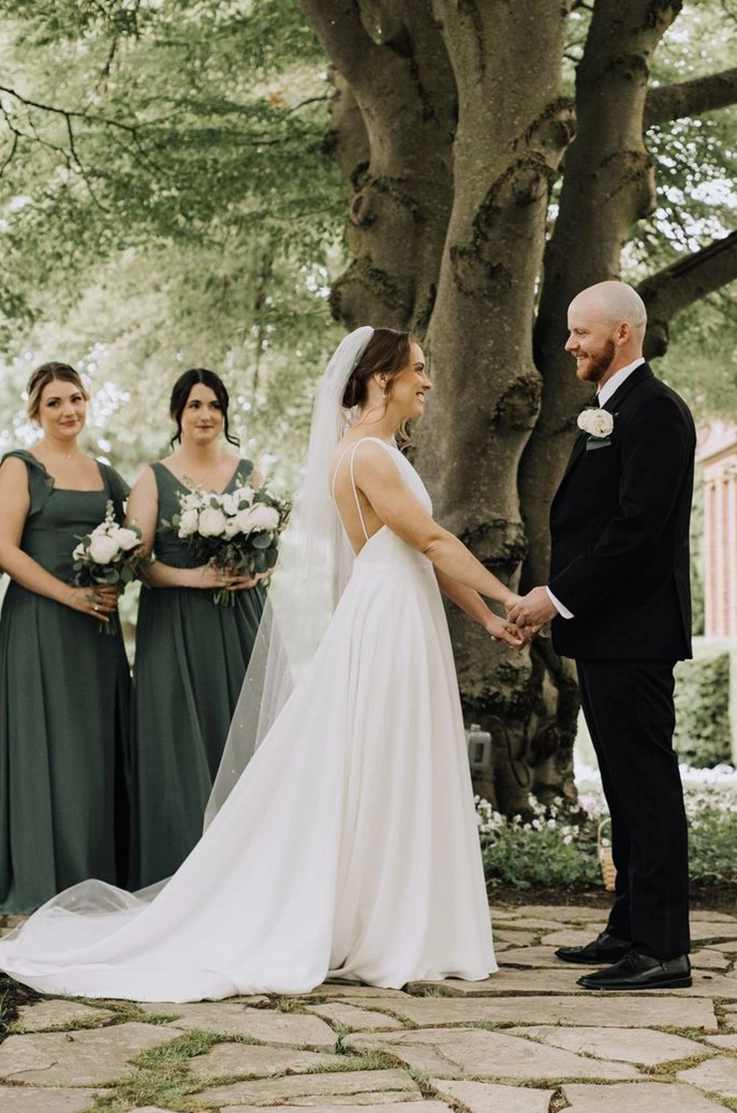 Bride in a spaghetti strap crepe wedding dress with a veil, faces her groom as they exchange vows at a large tree.