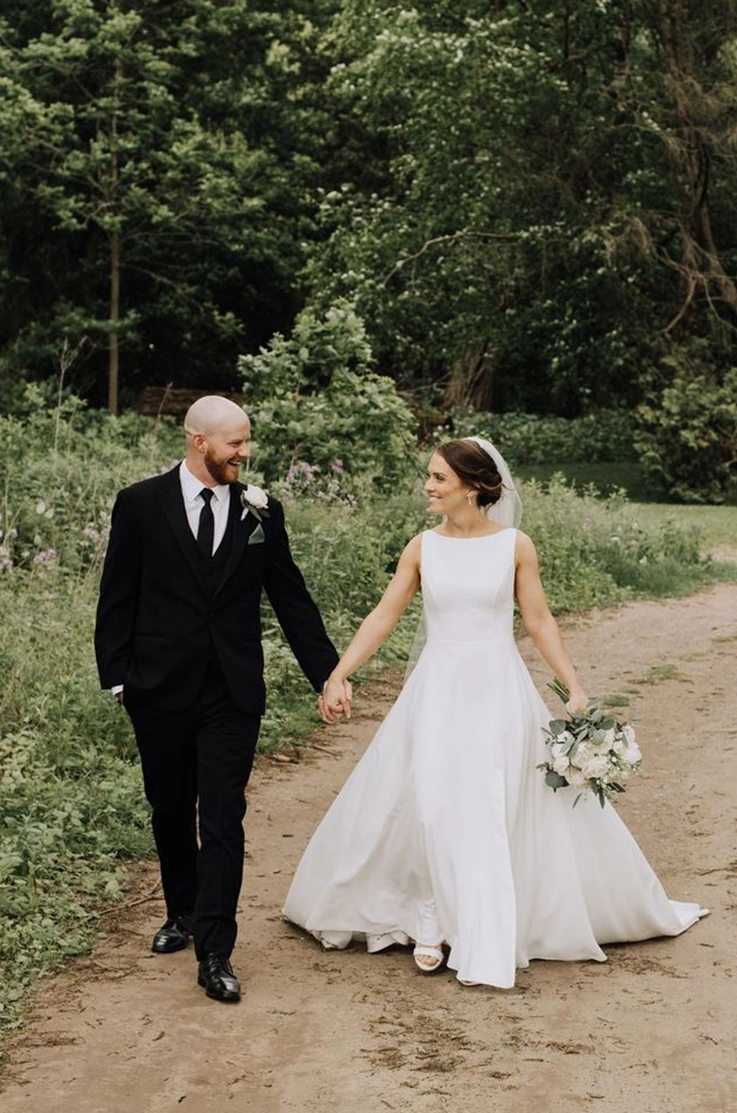 Bride in a bateau neck crepe gown with a veil, holds a bouquet and walks hand in hand with groom on an outdoor path.