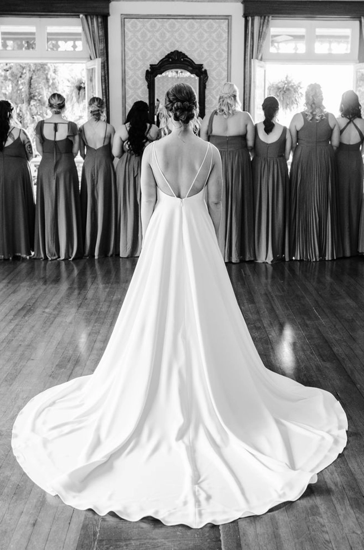 Back of bride in a crepe A-line wedding dress with back spaghetti straps, standing in front of bridesmaids, in black and white