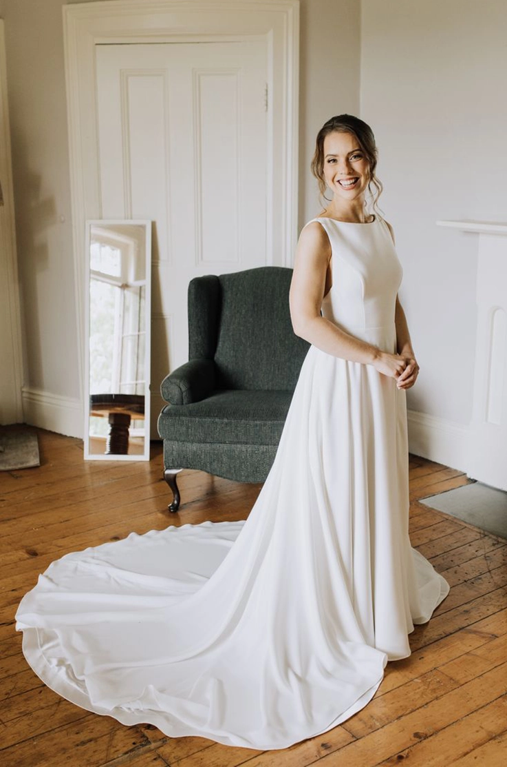 Smiling bride poses in a crepe sleeveless gown with a bateau neckline and full skirt, standing indoors.