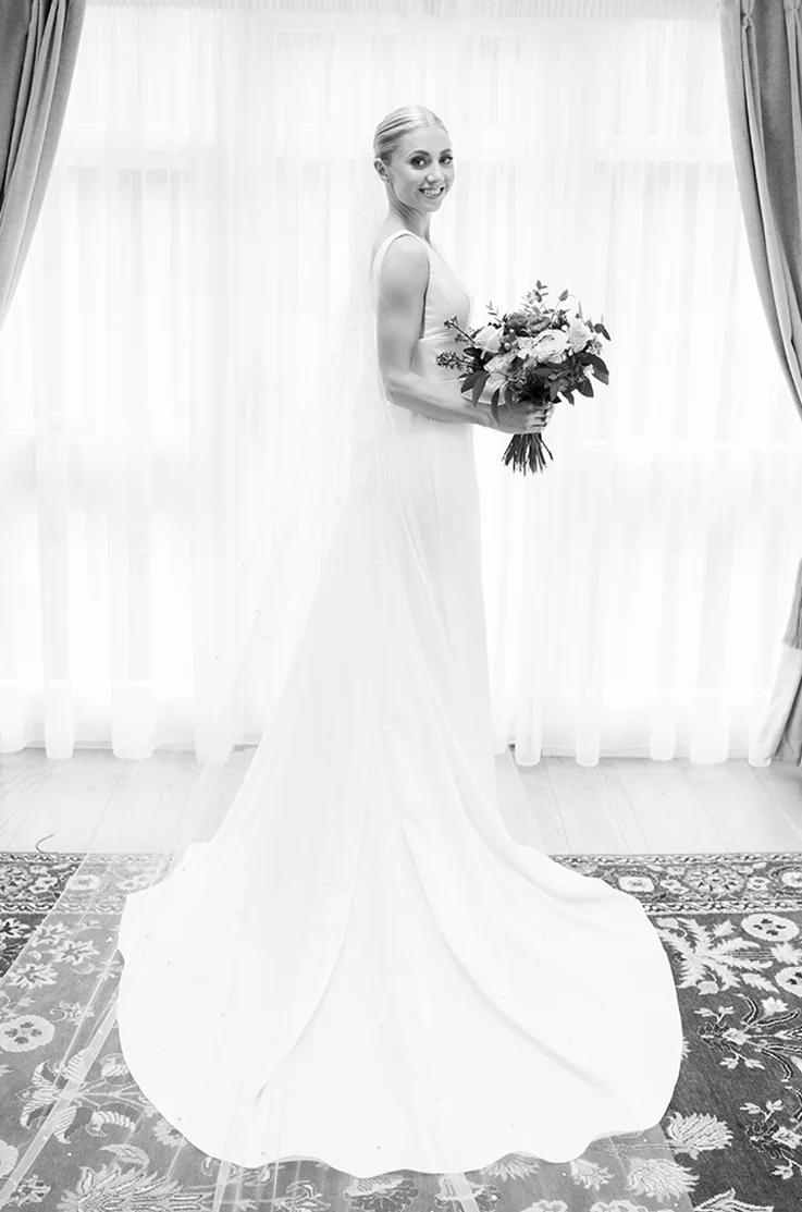Bride poses in a sleeveless satin gown with empire waist seam, holding a bouquet by a bright curtain, in black and white. 