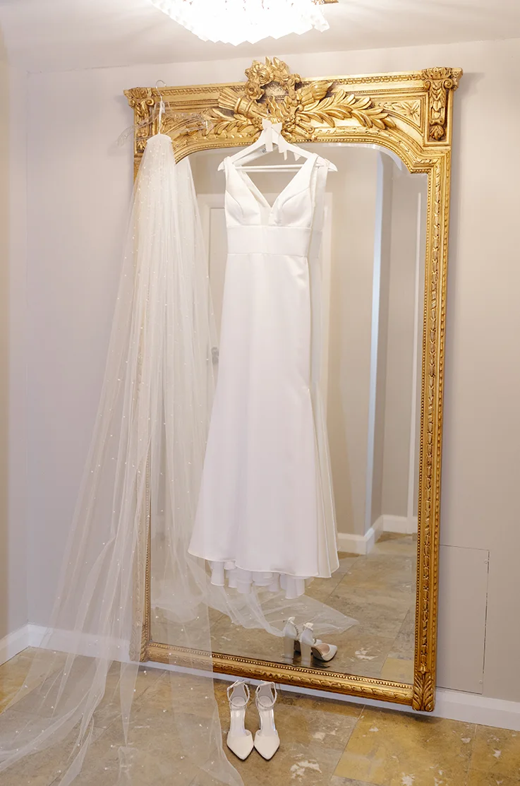 A sleeveless satin wedding dress with a V-neckline hangs on an ornate gold mirror next to a veil and shoes.