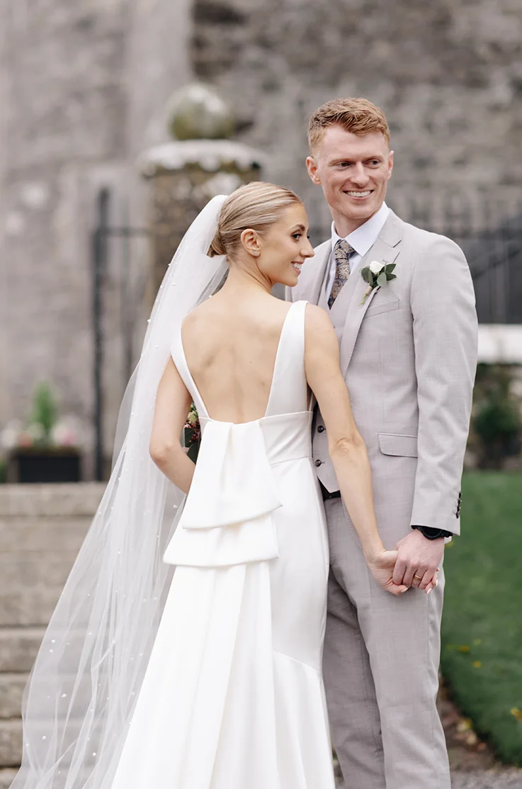 Back of bride in a satin backless gown with bow detail and a pearl-edge veil, posing alongside her groom outside the venue.