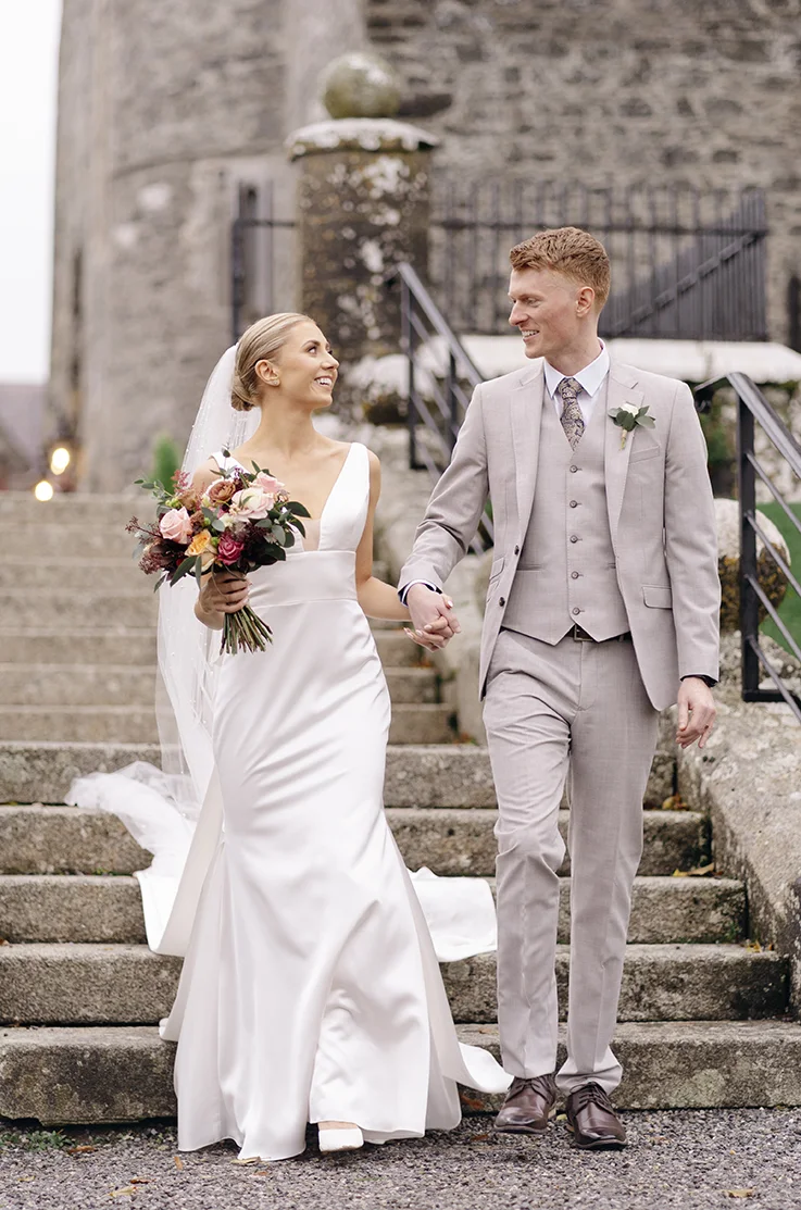 Bride in a plunging neck satin gown with an empire waist and veil, walks hand in hand with groom as they descend stone steps.