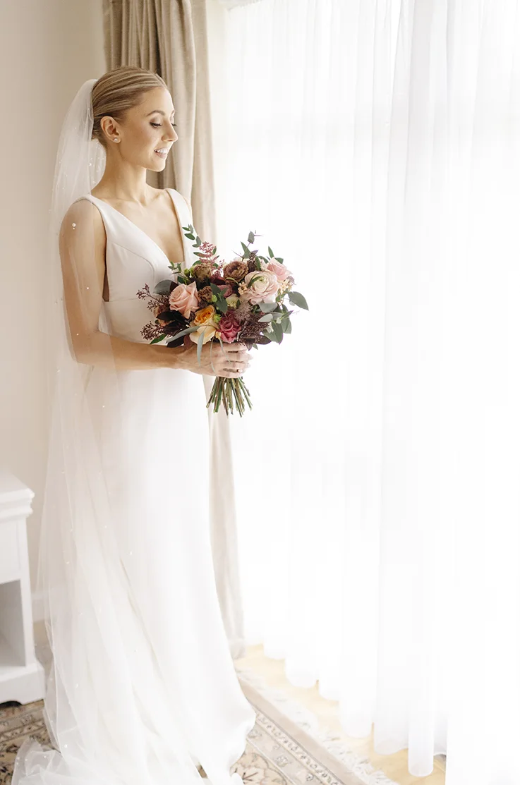 Bride poses by a bright window with curtains, in a V-neck satin gown with a pearl veil and empire waist indoors.